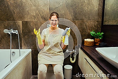 Angry woman sits on the toilet while cleaning the bathroom. Disinfection with yellow rubber gloves in the washroom Stock Photo