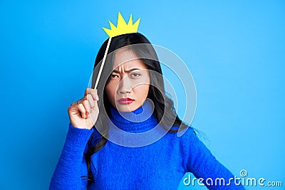 Angry young woman with paper crown on stick on blue background. Stock Photo
