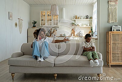 Annoyed Black mother sitting with offended son on sofa talking on phone. Parents and discipline Stock Photo