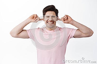 Annoyed young man shut ears with fingers and grimacing from annoying loud noise, noisy neighbours or bad music, standing Stock Photo