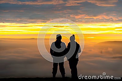 Anonymous couple standing on mountain together in love watching sunrise silhouetted stood high above clouds looking down valley Stock Photo