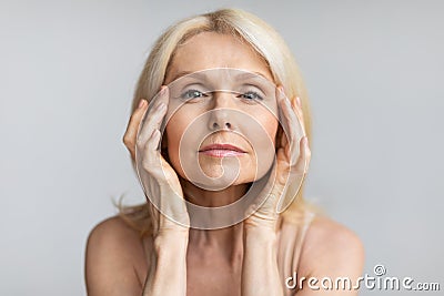 Antiage skincare. Senior woman touching skin near eyes and looking at camera, lady with flawless skin, grey background Stock Photo