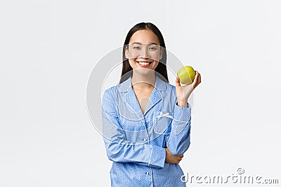 An apple keeps doctors away. Cheerful cute asian girl in blue pajamas eating apple for breakfast and smiling joyful Stock Photo