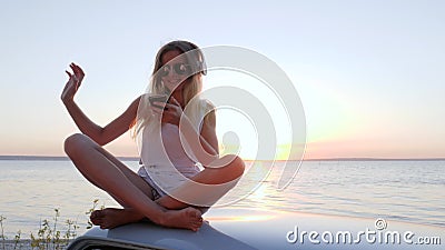 Appréciant la musique au toit de l'automobile sur la rivière de rivage, la fille avec des écouteurs de Bluetooth écoute des chans clips vidéos Appréciant la musique au toit de l'automobile sur la rivière de rivage, la fille avec des écouteurs de Bluetooth écoute des chans clips vidéos