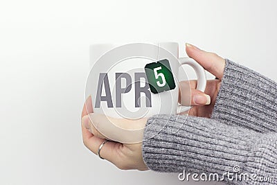 April 5th. Day 5 of month, Calendar date. Closeup of female hands in grey sweater holding cup of tea with month and calendar date Stock Photo