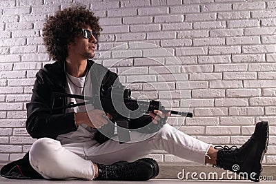 an Arab with an African hairstyle in stylish fashionable clothes with an airsoft gun poses against a brick wall Stock Photo