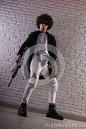 an Arab with an African hairstyle in stylish fashionable clothes with an airsoft gun poses against a brick wall Stock Photo