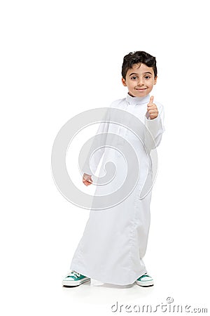 Arab boy with thumbs up and a smile, leaning to the side, wearing white traditional Saudi Thobe and sneakers, raising his hands on Stock Photo