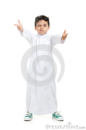 Arab boy winking and acting cool with crossed hands, wearing white traditional Saudi Thobe and sneakers, on white isolated Stock Photo