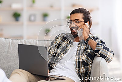 Arab guy sitting on sofa with laptop, having phone coversation Stock Photo