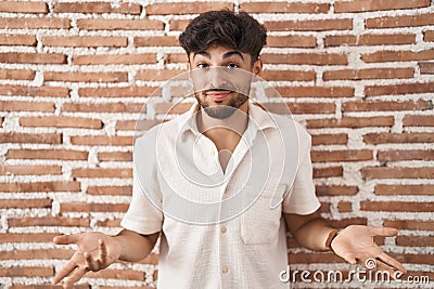 Arab man with beard standing over bricks wall background clueless and confused with open arms, no idea concept Stock Photo