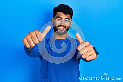 Arab man with beard wearing casual blue sweater approving doing positive gesture with hand, thumbs up smiling and happy for Stock Photo