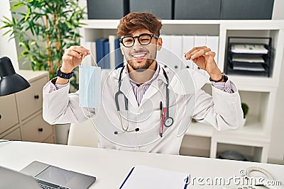Arab man with beard wearing doctor uniform holding medical mask winking looking at the camera with sexy expression, cheerful and Stock Photo