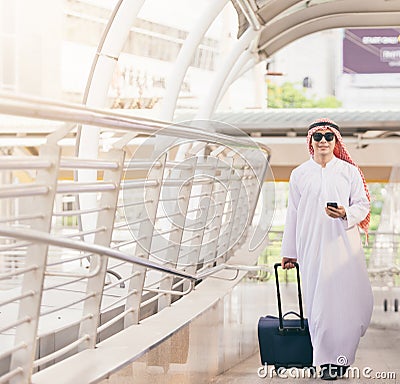 Arab man in travel concept . Young saudi arabic man in traditional clothes walking with suitcase on airport background. Stock Photo