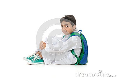 Arab school boy sitting on ground with a smile on his face, wearing white traditional Saudi Thobe, back pack and sneakers, raising Stock Photo