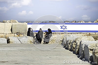 Arab women with Chador, sitting in the port of Old Jaffa with the Israeli flag in the background. The concept for peace. Editorial Stock Photo