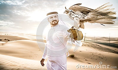 Arabic man with traditional emirates clothes walking in the desert with his falcon bird Stock Photo