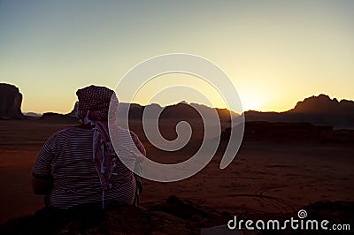 An arabic man wearing Jordanian keffiyeh head wrap is sitting on top of a rock overlooking the Wadi Rum Desert i Stock Photo