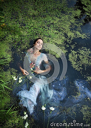 Art beautiful romantic woman lies in swamp in blue long dress with flowers. Portrait brunette in transparent dress in water swamp Stock Photo