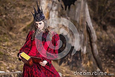 Art Photography. Mysterious Fairy Medieval Queen in Red Dress and Spiky Black Crown Posing With Ancient Book in Forest in Early Stock Photo