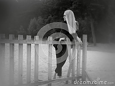 A black and white photograph of a girl, who stands on the sand on the beach, leaning on a broken wooden fence Stock Photo