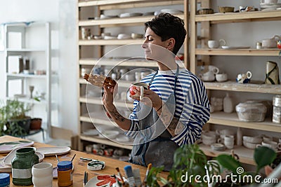 Artisan tattooed girl drink coffee and looking at tasty bun lustily in art studio during break Stock Photo