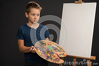 Artist little boy in blue t-shirt looking sad holding a palette with paints dried upstanding next to an canvas on the Stock Photo
