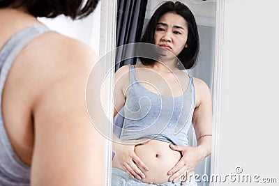 Asain woman checking her abdominal and looking at her fat belly in a mirror, weight gain concept Stock Photo