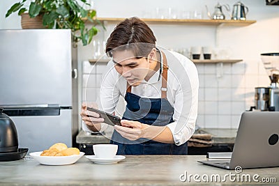 Asian businessman cafe owner take photo of cup of coffee for marketing. Young barista man restaurant worker use laptop computer Stock Photo