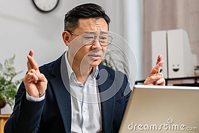 Asian businessman crossed fingers looking at laptop screen asking for good luck news win victory Stock Photo