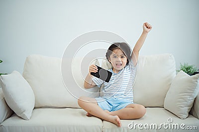 Asian child girl raise your hand cheerful success looking using and touch mobile phone screen on couch sofa. Baby smiling funny Stock Photo