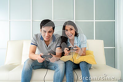 Asian couple man ans woman to playing video games with joysticks while sitting in sofa in living room at home, concept of family Stock Photo