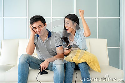 Asian couple man ans woman to playing video games with joysticks while sitting in sofa in living room at home and woman winning in Stock Photo