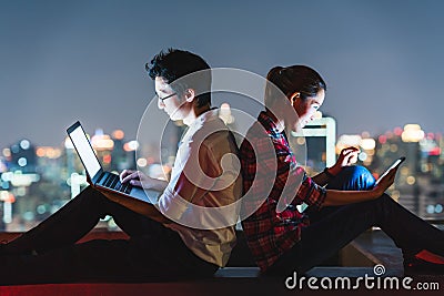 Asian couple using laptop and smartphone together, lean on each other on rooftop at night Stock Photo