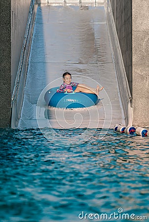 Asian cute child girl enjoying summer vacation, riding blue float ring sliding from water ramp to swimming pool, vertical image Stock Photo