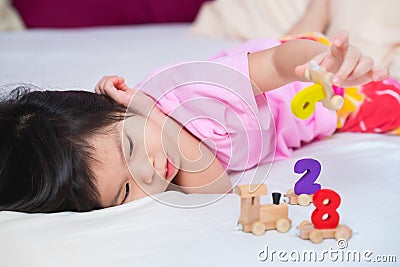 Asian cute girl lying playing wooden toys on a soft white mattress. Adorable kid play number train. Little child`s relax moment. Stock Photo