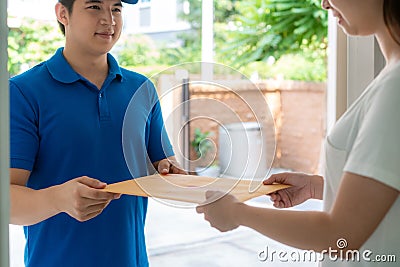 Asian delivery young man in blue uniform smile and holding letter or document envelope in front house and Asian woman accepting a Stock Photo