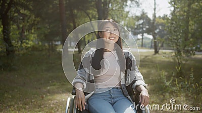 An asian disabled female with long hair is posing, sitting in a wheelchair in a public park, smiling and touching hair Stock Photo