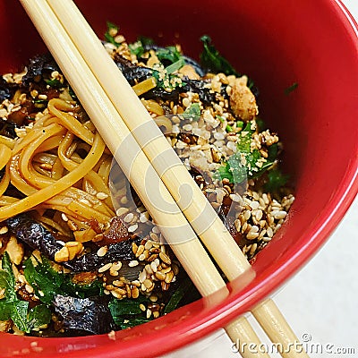 Asian dish in a red bowl, wooden sticks, sesame and small pieces of algae, long noodles and peanuts, Korean food, Asian dish Stock Photo
