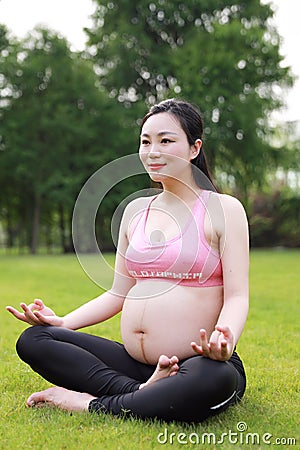 Asian Eastern Chinese pregnant woman do yoga sit in meditation in nature outdoor on grass meadow do lotus pose keep fit healthy Stock Photo