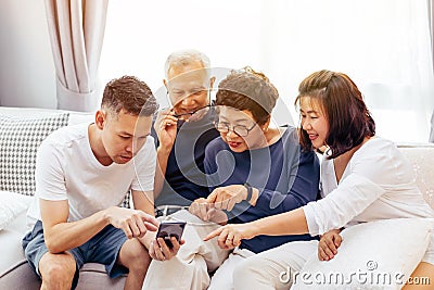 Asian family with adult children and senior parents using a mobile phone and relaxing on a sofa at home together. Stock Photo