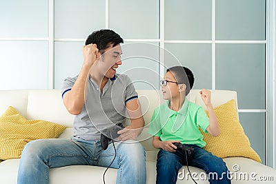 Asian family father and his son to playing video games with joysticks and arm raised to winning in game while sitting in sofa in Stock Photo