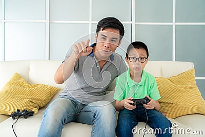 Asian family father teaching his son to playing video games with joysticks while sitting in sofa in living room at home, concept Stock Photo