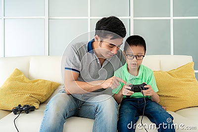 Asian family father teaching his son to playing video games with joysticks while sitting in sofa in living room at home, concept Stock Photo