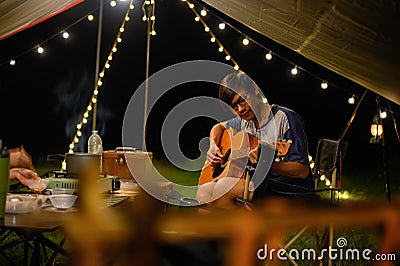 Asian family and friends vacation camp, Dinner and relaxing guitar play under tarp or flysheet, Outdoor camping on the courtyard Stock Photo