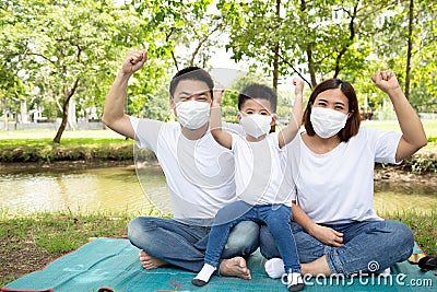 Asian family wearing protective medical mask for prevent virus Covid-19 and hand up and sitting together on floor at green public Stock Photo