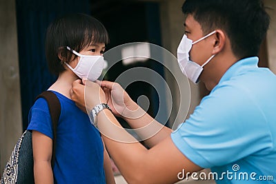 Asian father wearing protective mask puts protective cloth face mask on his little child, schoolkid with backpack is ready to Stock Photo