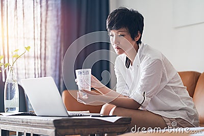 Asian female freelancer sitting on couch thinking & working on laptop at home Stock Photo