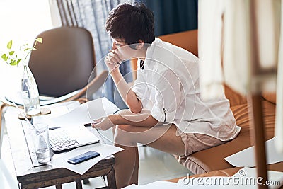 Asian female freelancer sitting on couch thinking & working on laptop at home Stock Photo