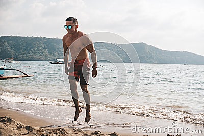 Asian fitness guy doing jumping jacks, star jump or burpees exercise at seaside outdoors. Healthy lifestyle by the beach Stock Photo